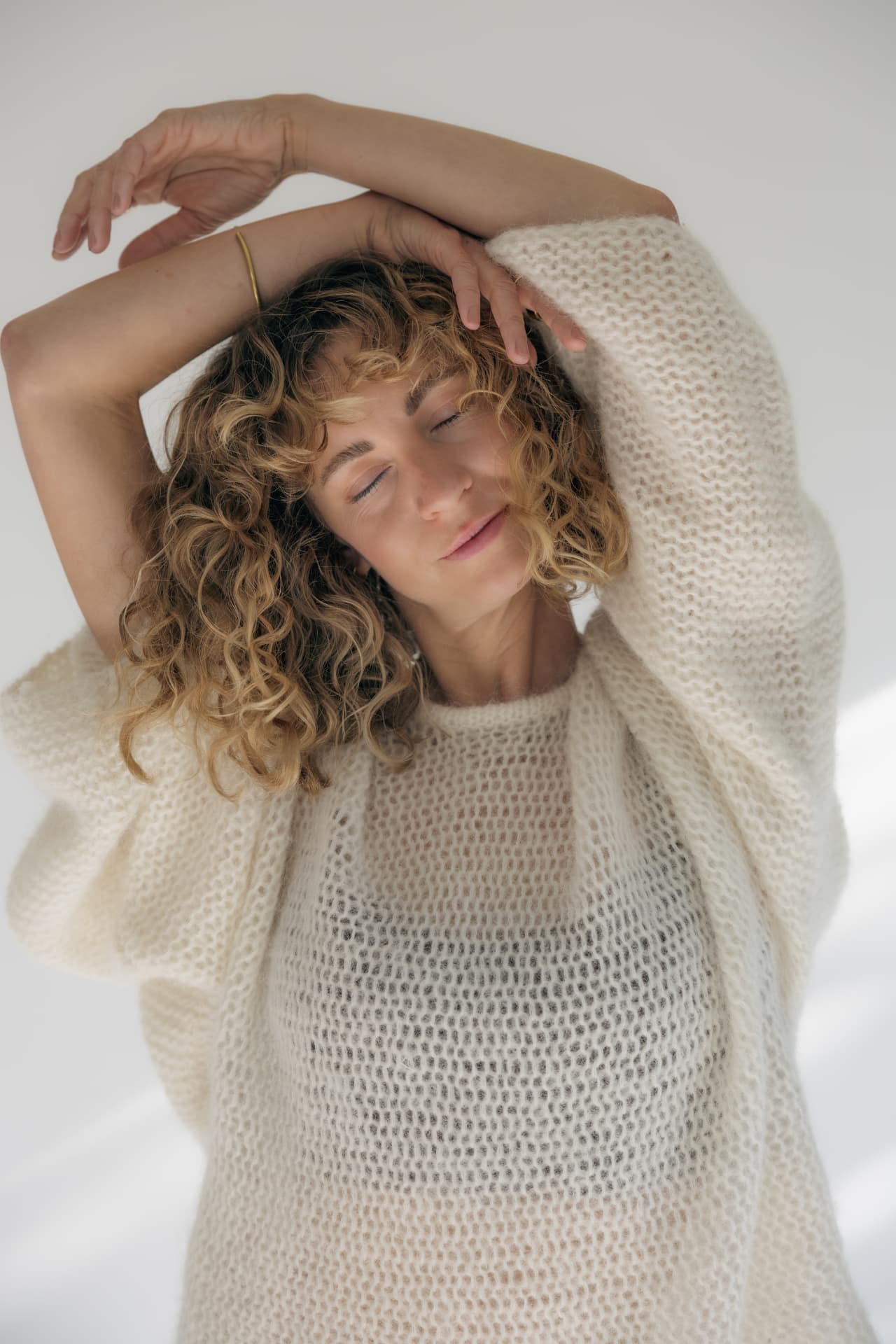 Woman with curly hair in a cream knit sweater, arms folded above her head