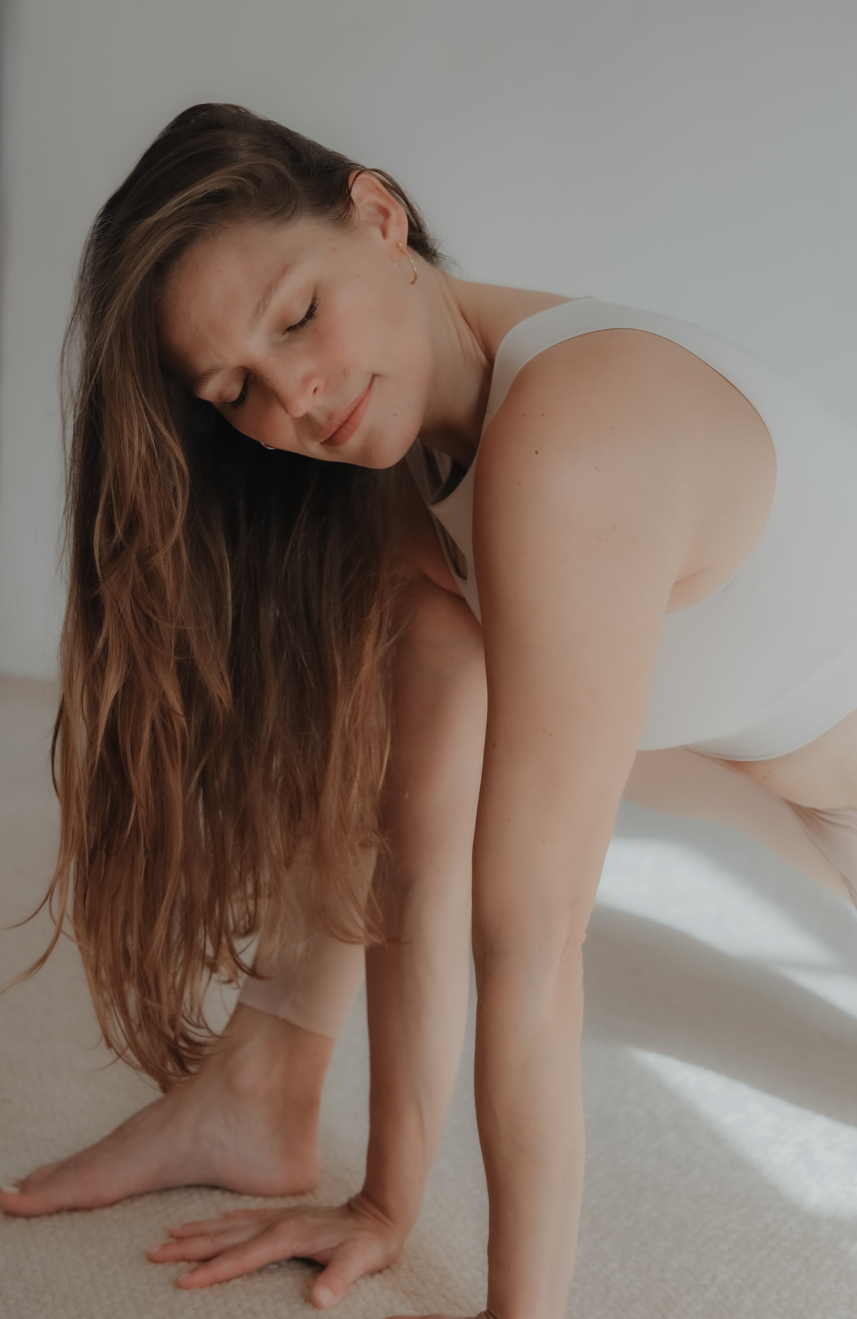 Woman in a low lunge stretch on a pale carpet, one hand planted, eyes closed in soft daylight