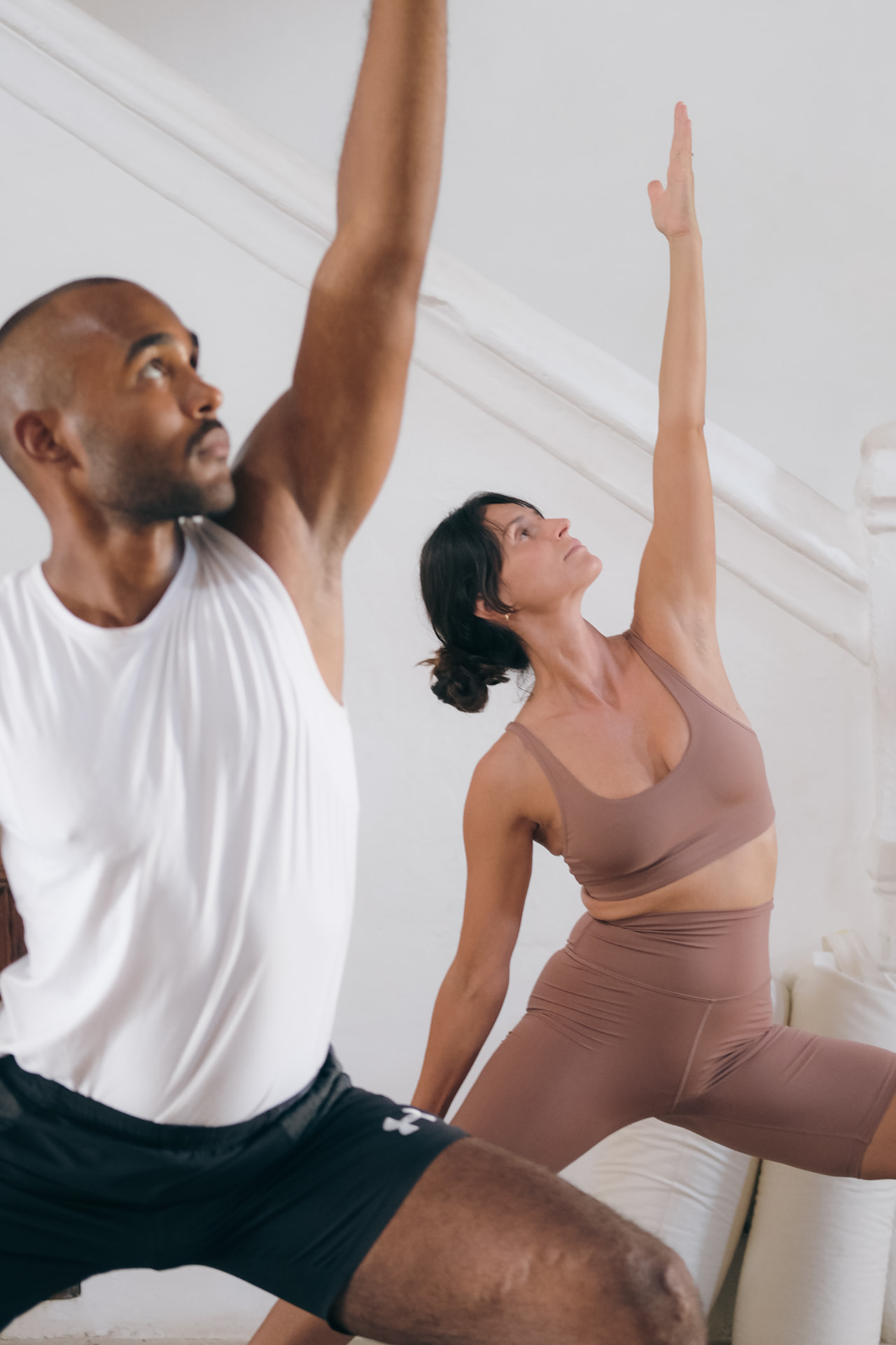 Two people in a wide lunge with one arm reaching toward the ceiling in a bright white studio