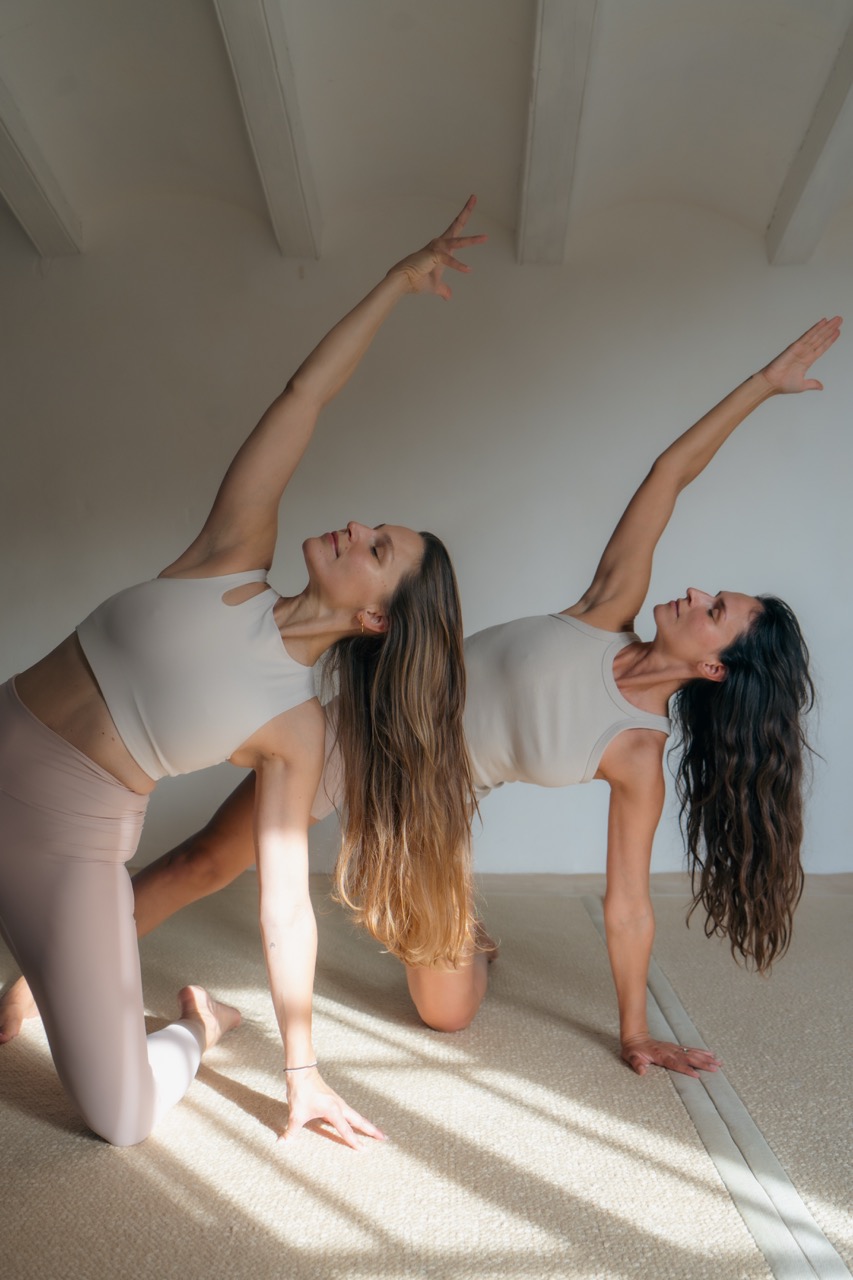 Two women in matching neutral activewear kneeling in a flowing dance posture, one arm stretched overhead