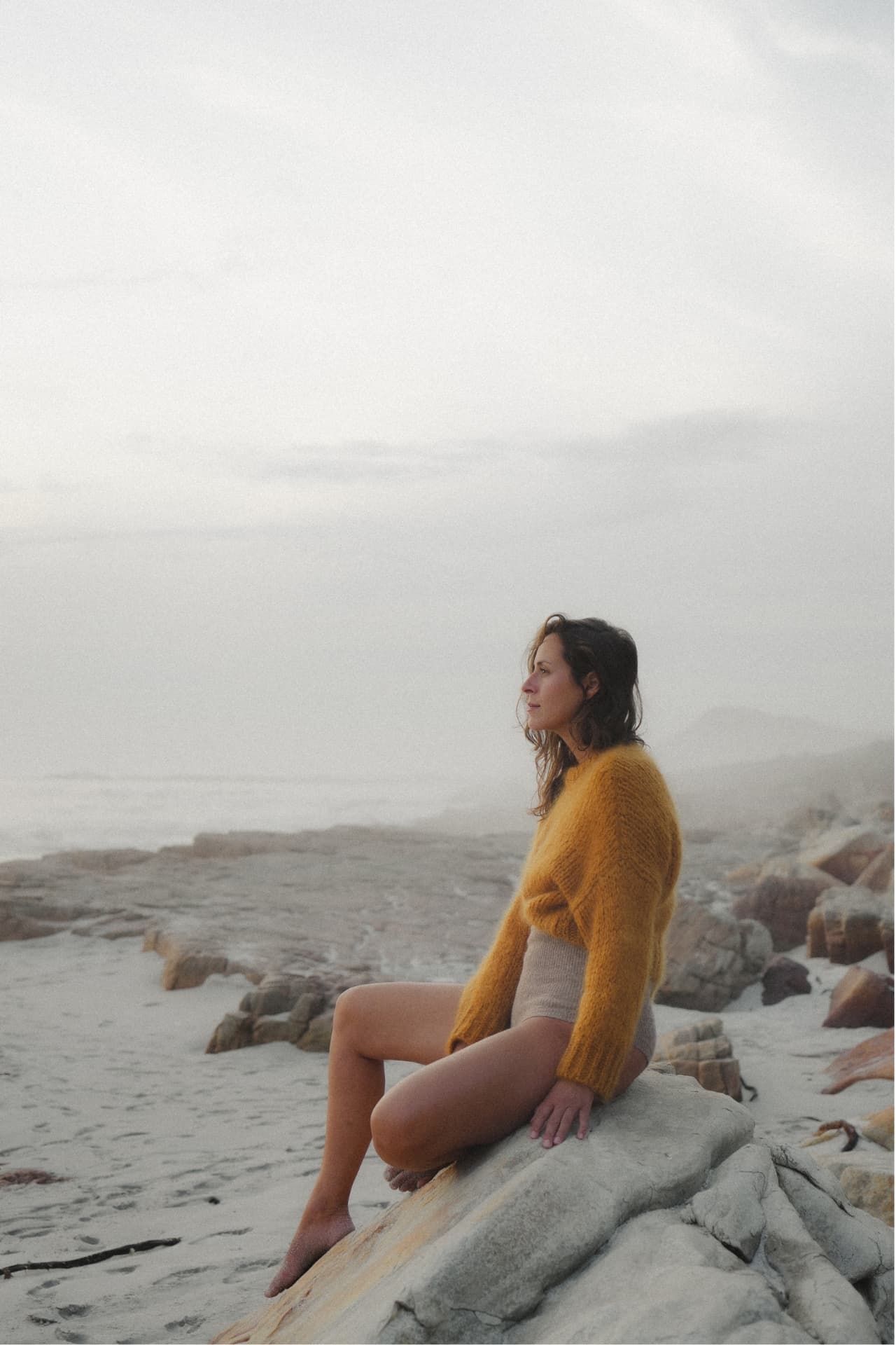 Julie Zimmermann sitting on a coastal rock in sea fog, wearing a mustard knit sweater