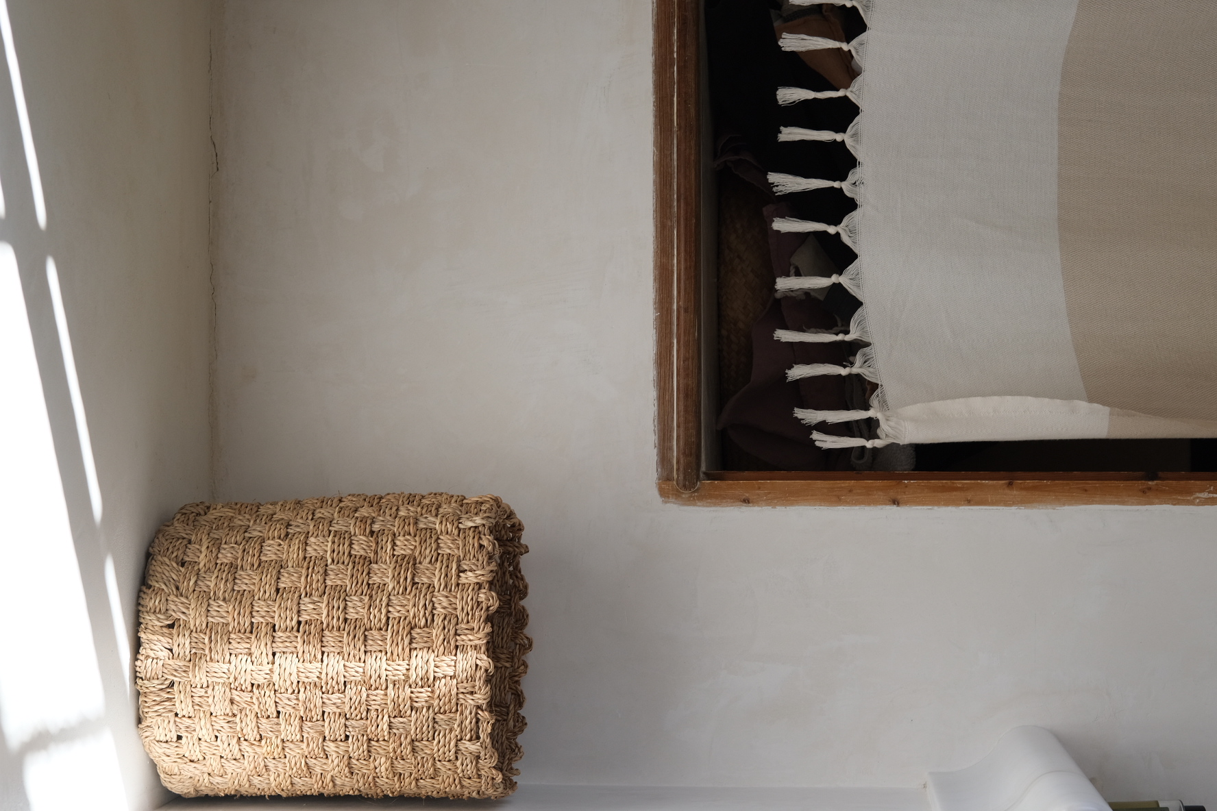Chunky woven straw basket beside a wooden opening draped with a fringed cream throw in strong daylight