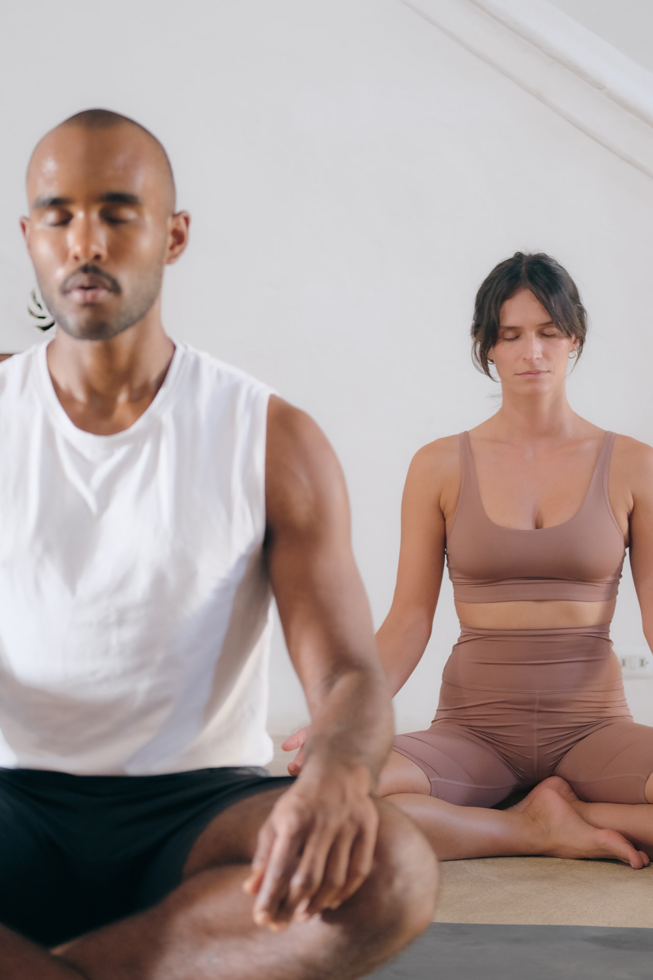Two people seated cross-legged in meditation, eyes closed, in a plain bright room