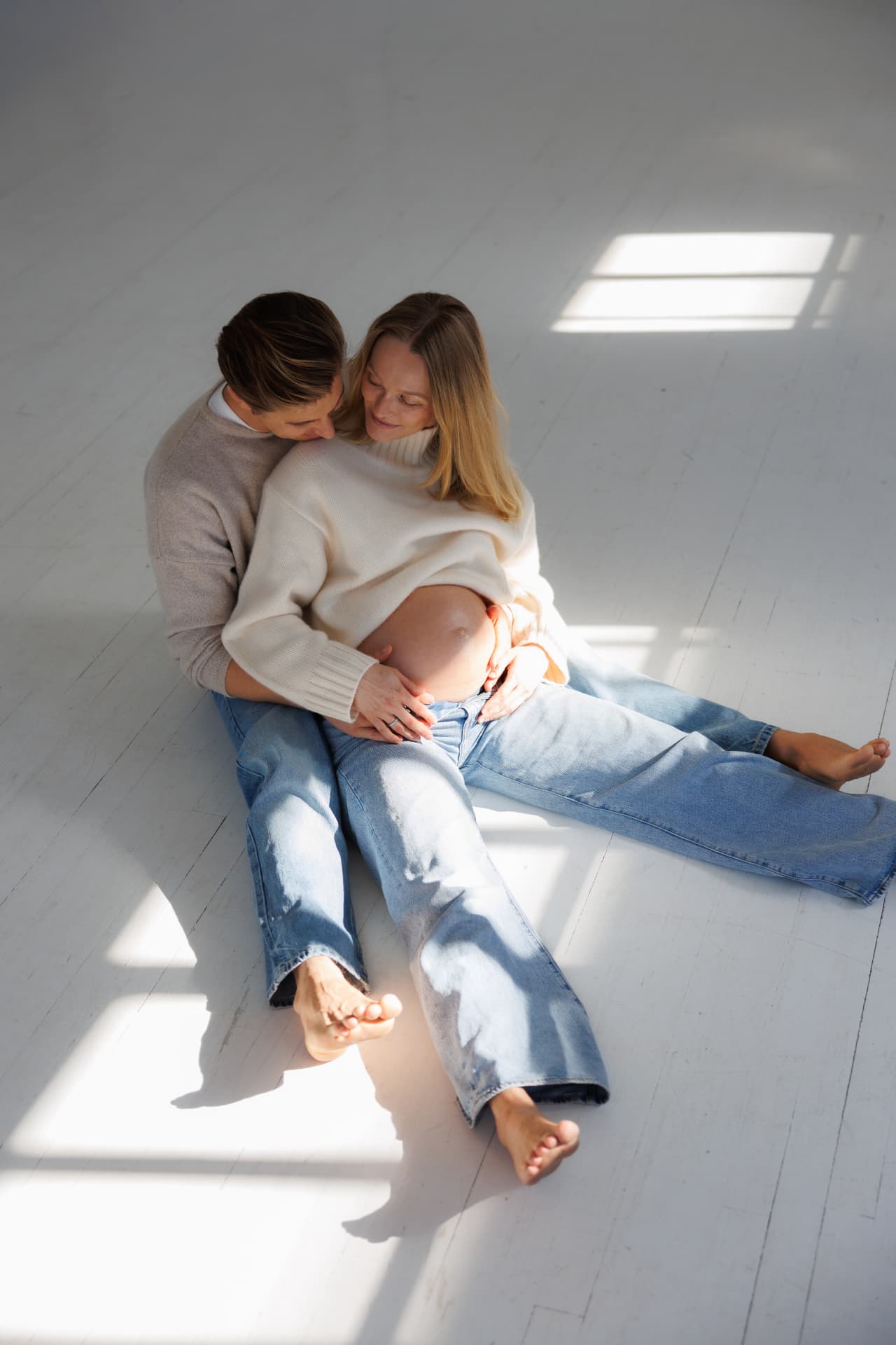 Expectant parents seated together on a white wooden floor in soft window light
