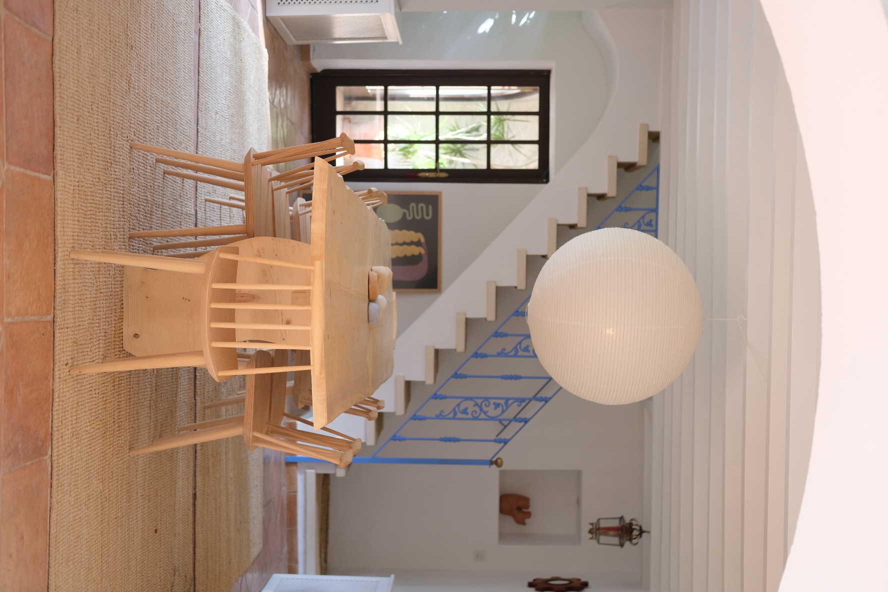 Dining area with a light-wood table and chairs, a blue-railed staircase and a spherical paper pendant lamp